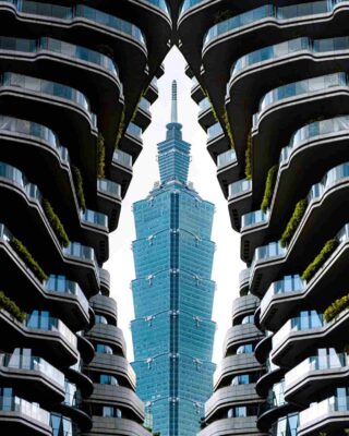 A striking architectural shot of Taipei 101 towering over the surrounding buildings in the Xinyi district, framed by the modern, terraced balconies of a nearby structure.
