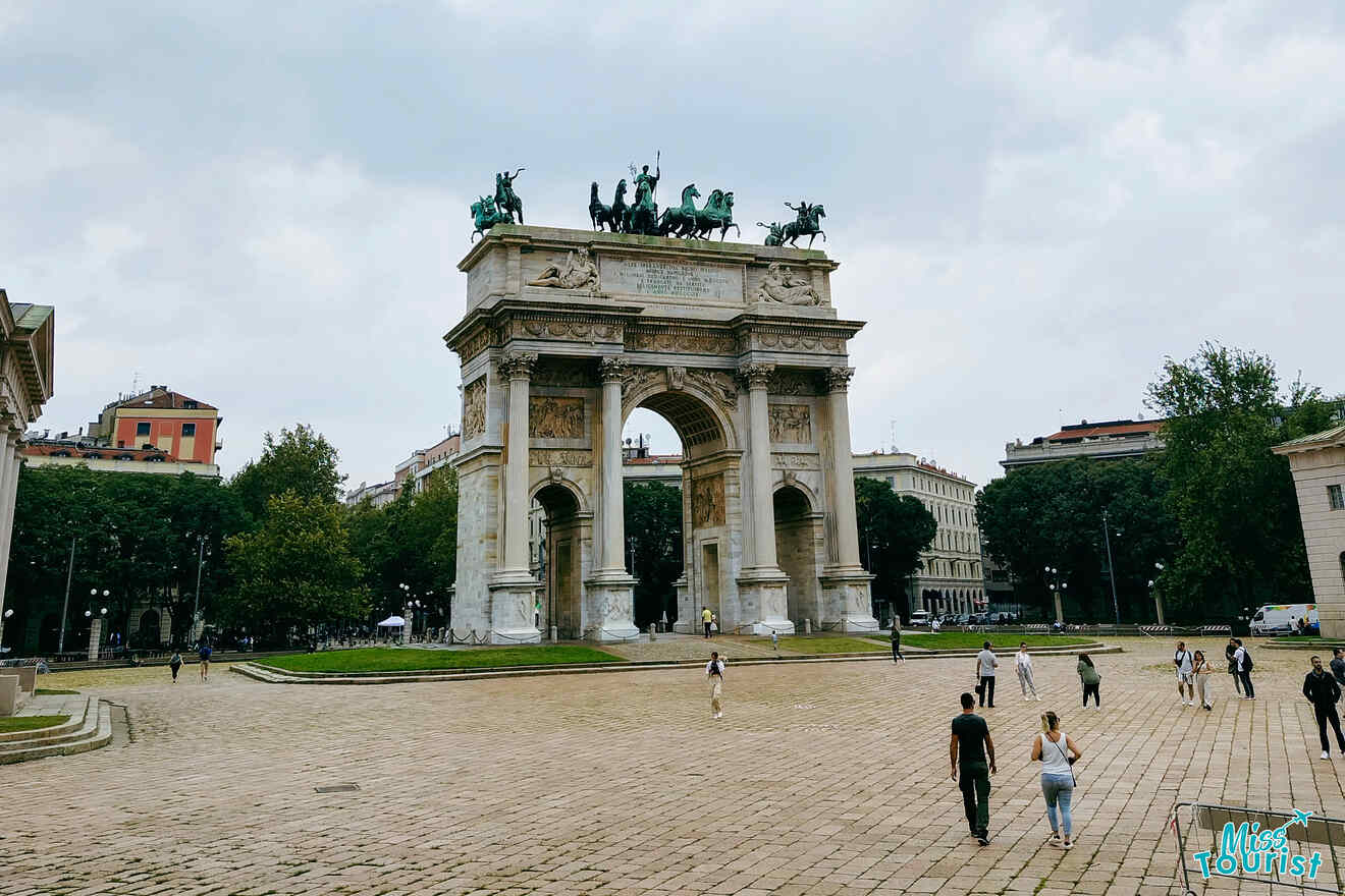 The neoclassical Arch of Peace (Arco della Pace) in Milan, with sculptures atop, viewed from a cobblestone plaza with pedestrians under a cloudy sky