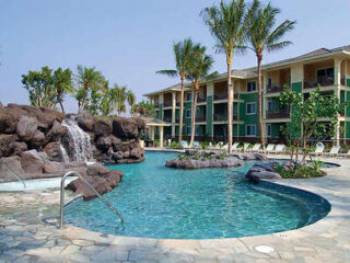 Resort-style pool with palm trees, natural rock formations, and a waterfall, framed by a multi-story hotel building