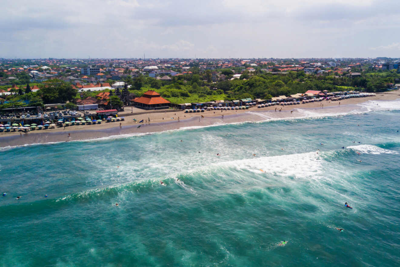 Aerial view of a vibrant beach in Canggu with surfers riding the waves, beach umbrellas lining the shore, and a bustling street scene alongside the coast