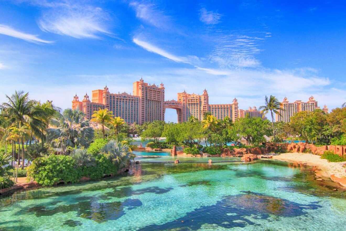 Majestic view of a grand pink resort with multiple towers, set amidst lush tropical vegetation, overlooking a clear blue lagoon under a sunny sky with wispy clouds.