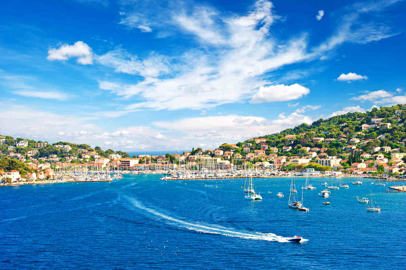 A vibrant view of the harbor in Saint-Tropez, featuring a bustling marina with moored yachts, flanked by the lush green hills and colorful houses of the town, beneath a bright blue sky scattered with white clouds