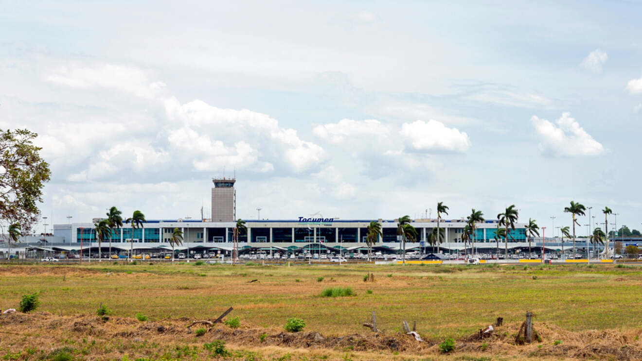 Exterior view of Tocumen International Airport in Panama, showcasing the busy terminal with parked cars and palm trees under a partly cloudy sky