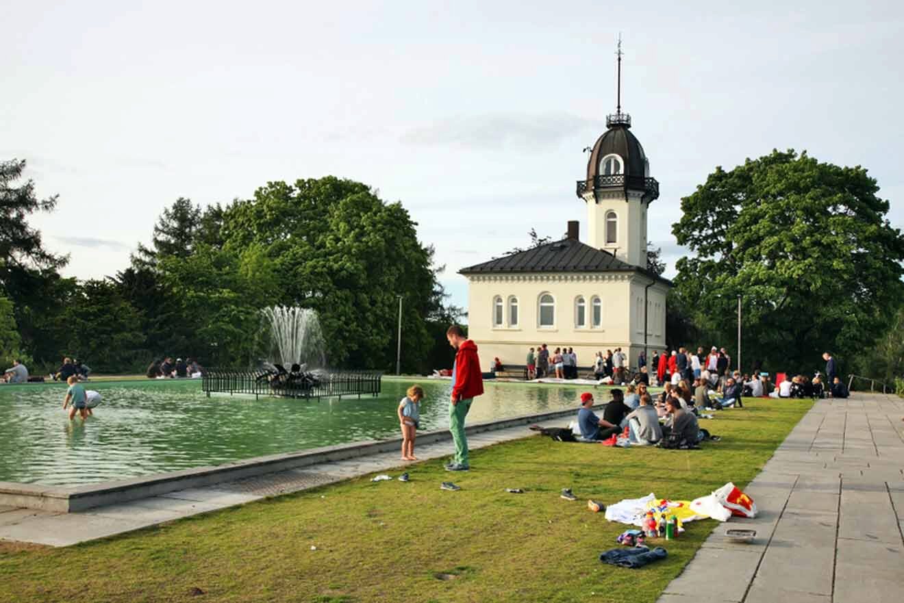 Public park scene with people relaxing by a fountain and pond, with a classic white building featuring a clock tower in the background, amidst lush green trees.