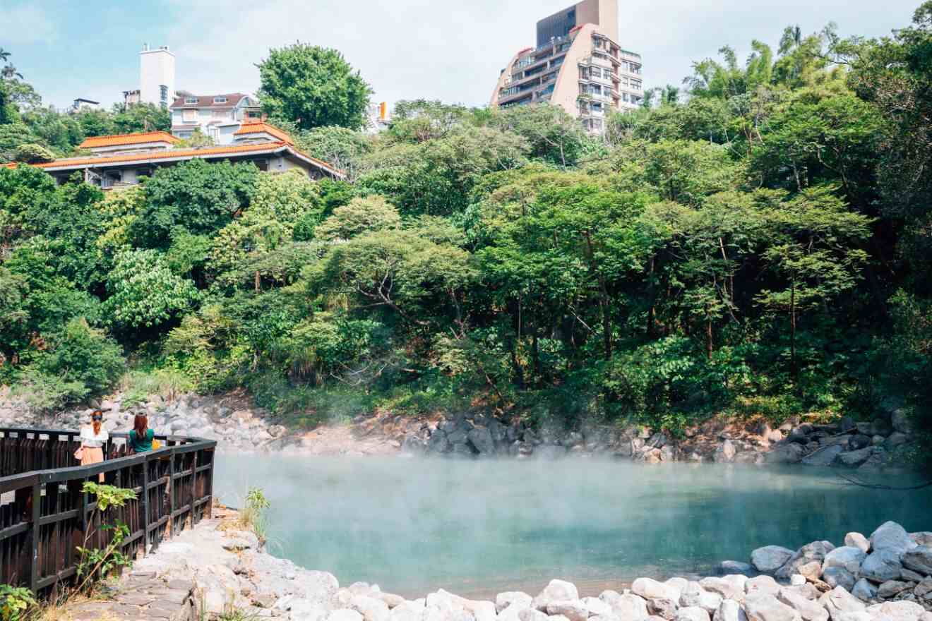 Hot spring with steam rising above clear blue water beside a wooden walkway, with lush greenery and urban buildings in the background
