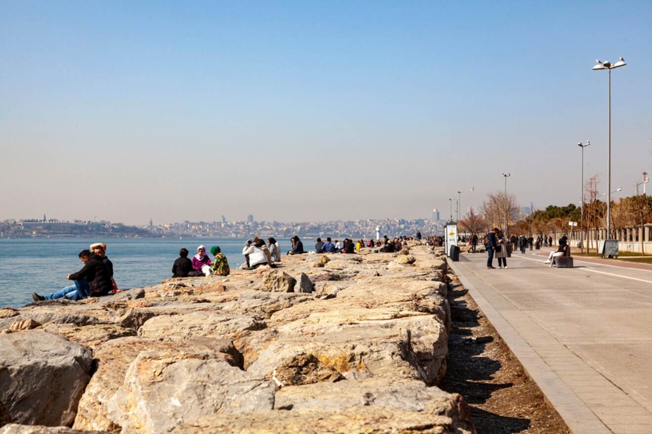 A seaside promenade in Istanbul with people sitting on rocks and walking along the path, enjoying a clear day with a view of the city across the water