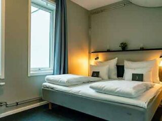 Minimalist hotel room with a comfortable double bed, soft lighting, and a large window, promising a restful stay for visitors.