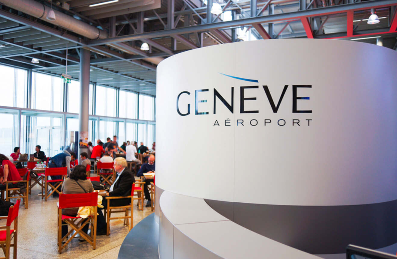 Inside Geneva Airport, with travelers sitting at tables in a dining area, and a prominent 'GENEVE AÉROPORT' sign