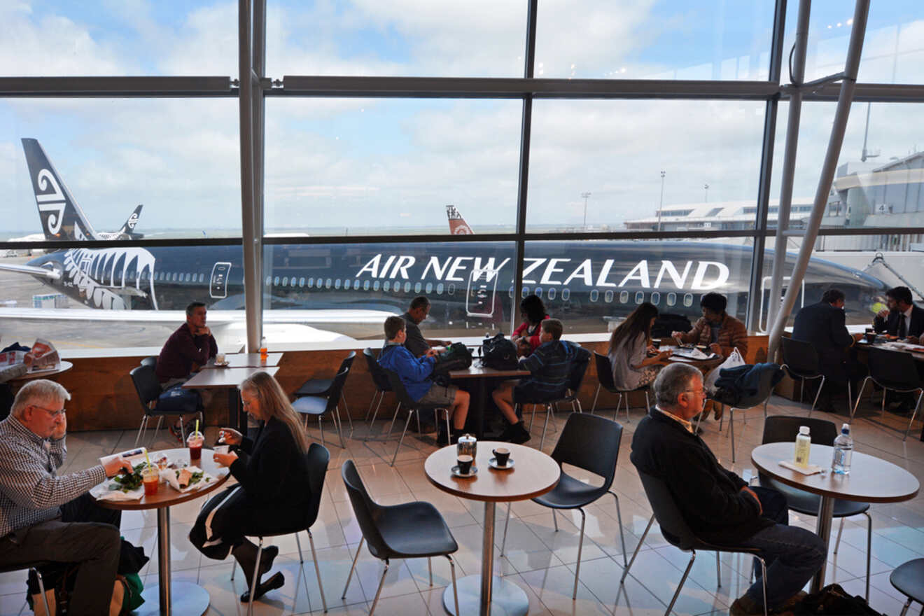 Airport lounge with travelers seated at tables, enjoying meals and conversations, with a large window view of an Air New Zealand airplane on the tarmac