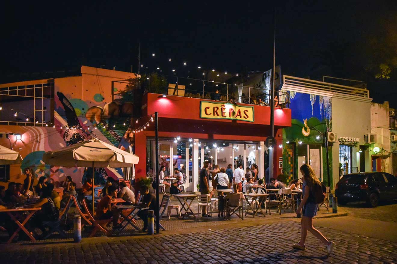 A bustling outdoor dining scene at night with string lights above, patrons seated at picnic tables outside a vibrant restaurant named 'CRÊPAS,' with a colorful mural on an adjacent building. A lone pedestrian walks by on the cobblestone street in Buenos Aires.