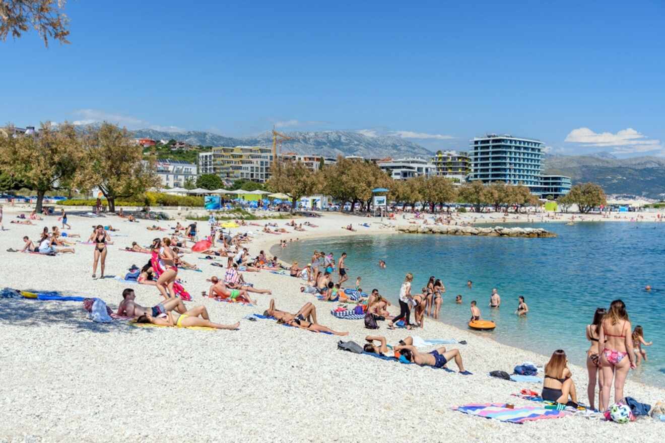 Crowded pebble beach scene at Znjan Beach in Split, with sunbathers and swimmers enjoying the clear blue waters with modern buildings and mountains in the distance