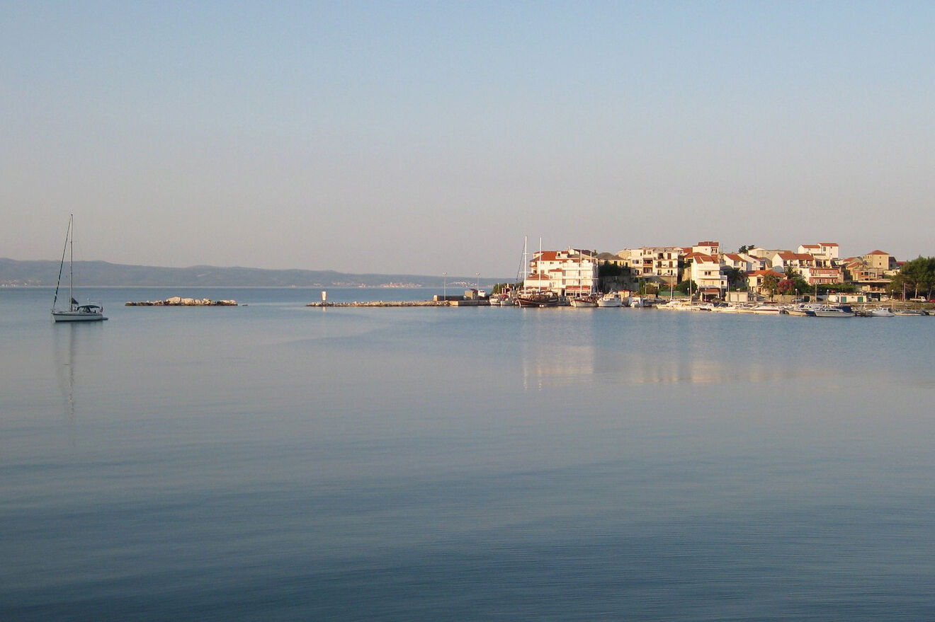 Tranquil early morning waters of Stobrec near Split, Croatia, with a solitary sailboat and a calm sea reflecting the sky, bordered by a sleepy coastal town awakening to daylight
