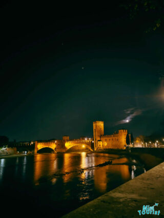 A nighttime scene of the Castelvecchio and Ponte Scaligero in Verona, Italy, with reflections on the Adige River and a clear night sky