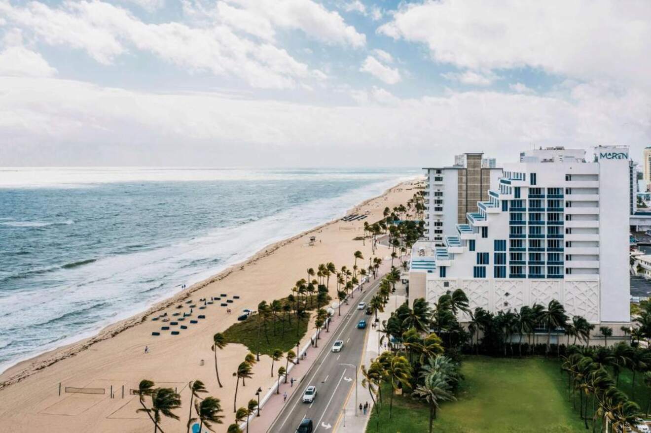 An oceanfront view captures a serene beach alongside a coastal road, with a modern hotel and palm trees framing the sandy shores