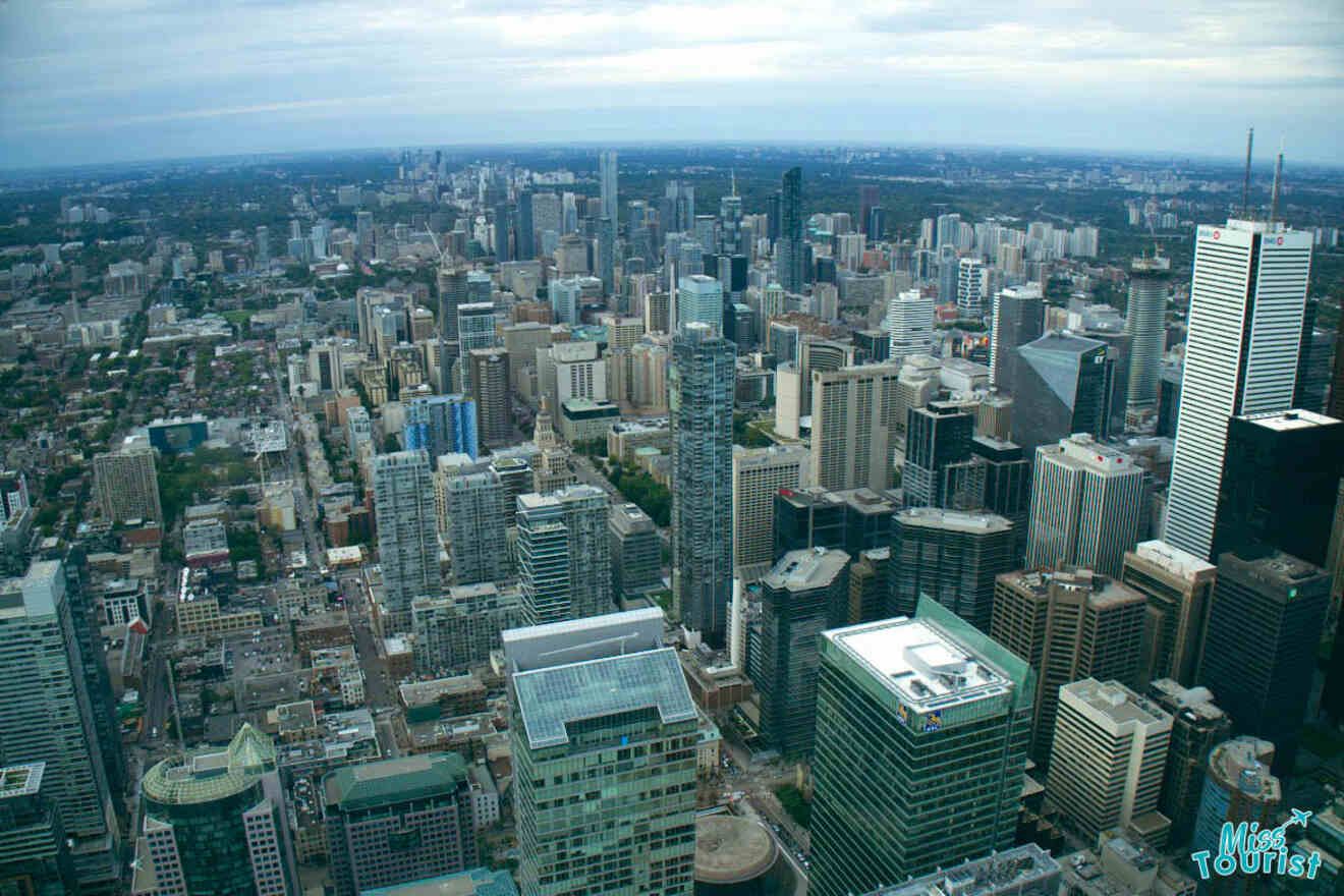 A daytime aerial view of Toronto's downtown, featuring dense high-rise buildings and the bustling cityscape extending to the horizon