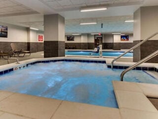 Indoor pool area with a hot tub in the foreground, metal handrails, lounge chairs, and wall art visible in the background.