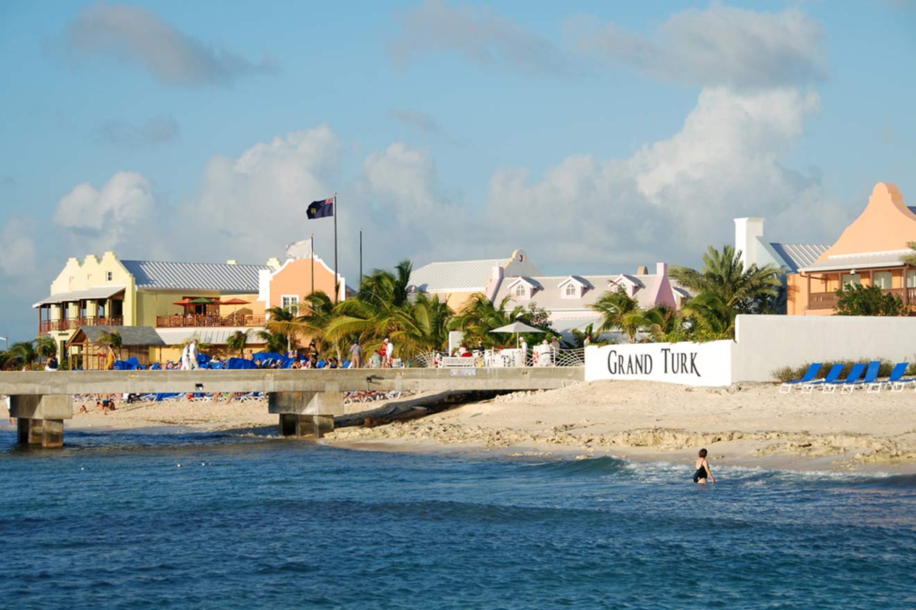 Seaside view of Grand Turk with colorful beachfront properties, blue beach chairs, and a prominent 'GRAND TURK' sign under a clear blue sky