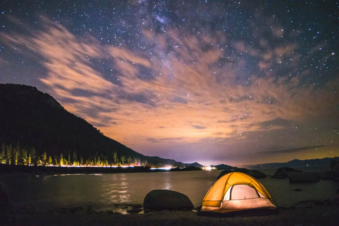 An illuminated tent glows under a starry night sky at Lake Tahoe, with a backdrop of silhouetted mountains and a shoreline dotted with lights from RVs and campgrounds.