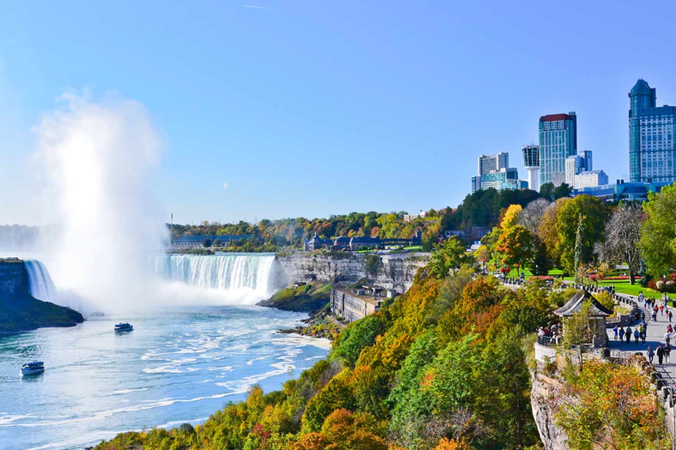 View of Niagara Falls with a misty haze rising from the cascade, surrounded by lush autumn trees, against a backdrop of the Niagara skyline on a clear day.