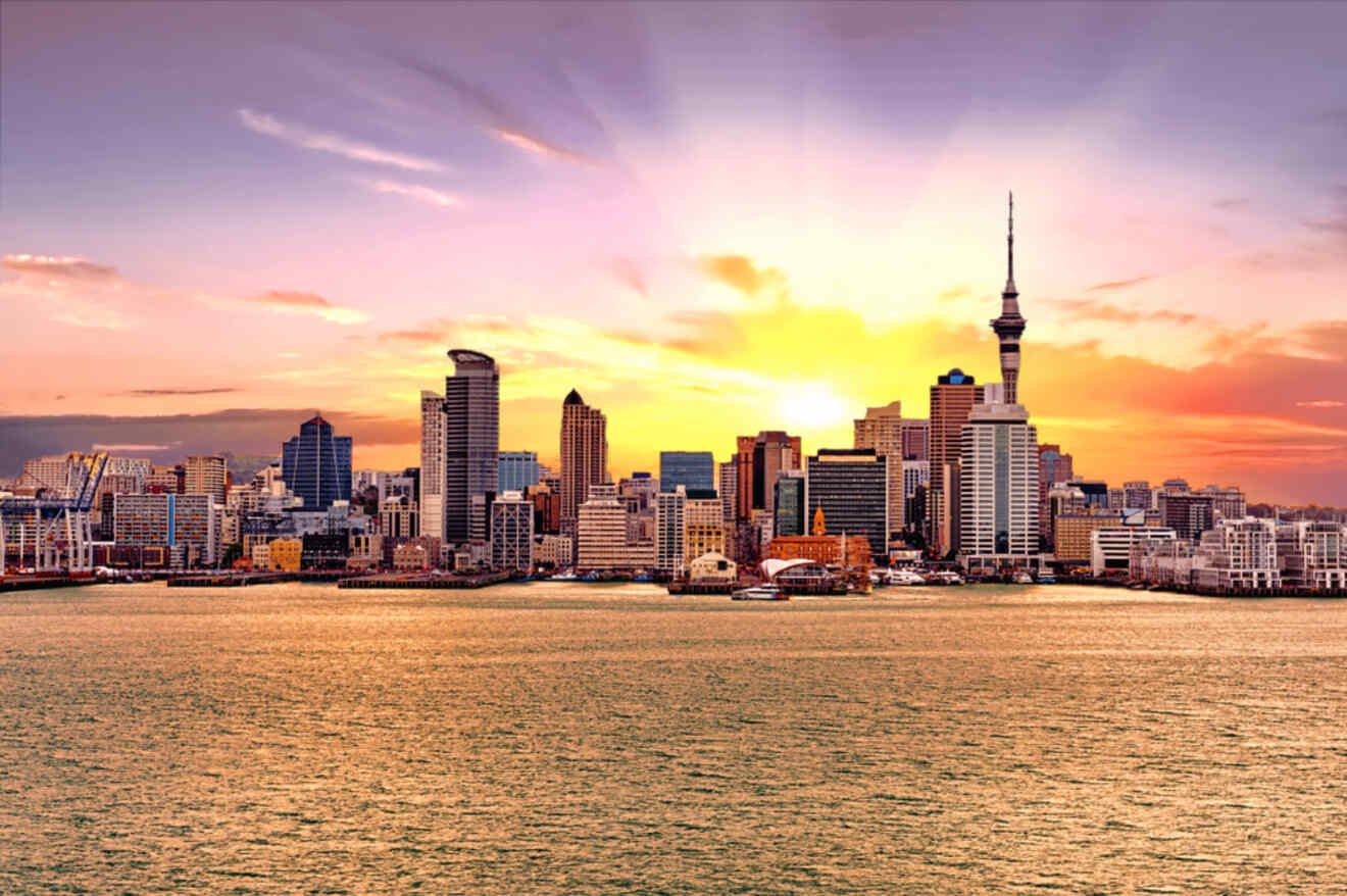 Skyline of Auckland at sunset, with the iconic Sky Tower standing tall amid the city's modern buildings, reflecting the warm glow of the setting sun over the waterfront