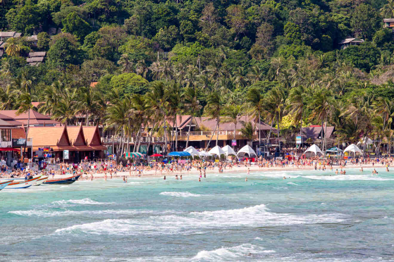 Crowded tropical beach with tourists and palm trees.