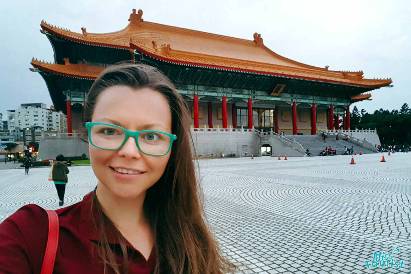 Selfie of the writer of the post with teal-framed glasses in the foreground, with the iconic National Theater in Taipei's Liberty Square visible in the background during daylight
