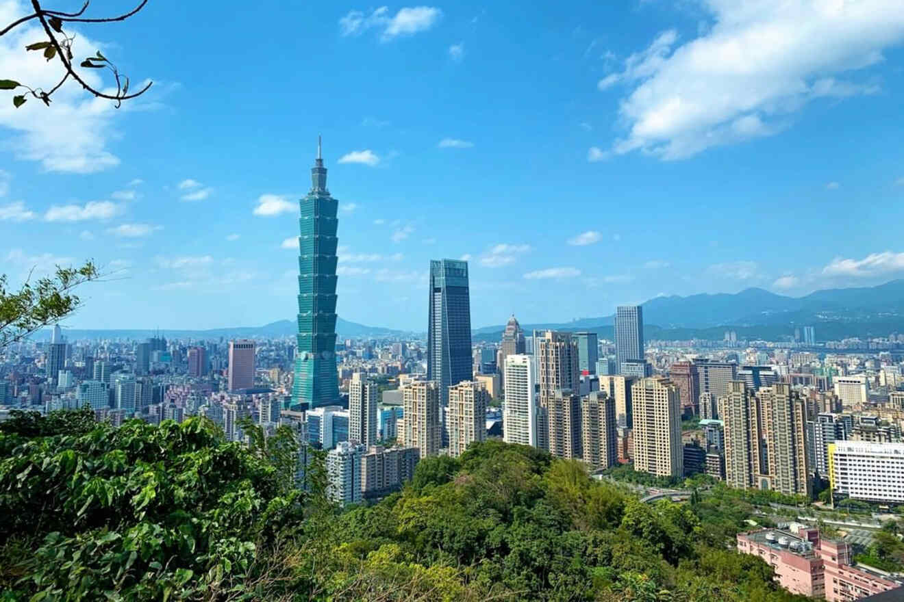 View of Taipei skyline featuring the iconic Taipei 101 skyscraper, surrounded by other high-rise buildings under a clear blue sky, with lush green foliage in the foreground