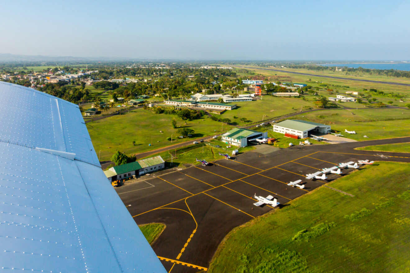 Aerial view of a small regional airport with multiple light aircraft parked on the tarmac, adjacent to airport buildings and hangars, with a clear blue sky overhead and a lush green landscape in the background.