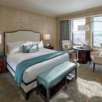A luxurious and well-lit hotel room with a large bed adorned with teal and white bedding, complemented by a richly textured carpet and a city view from the window.