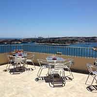A sunny hotel rooftop terrace with multiple dining tables overlooking a tranquil blue water body.