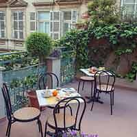 A cozy balcony setting with two metal chairs and a table, set with breakfast items, overlooking a view of neighboring European-style buildings and lush greenery.