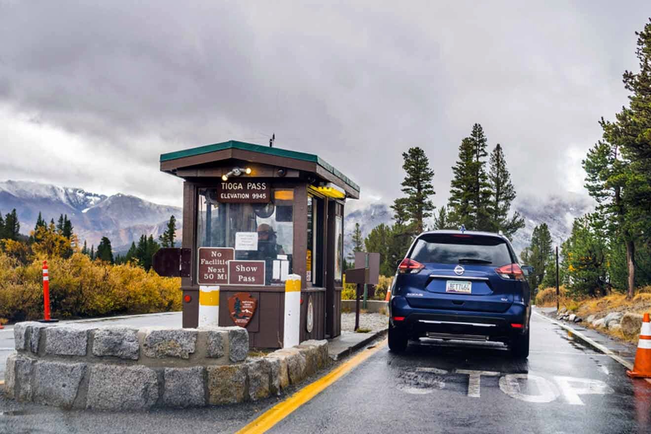 A vehicle stopping at Tioga Pass entrance booth in Yosemite under an overcast sky with snow-capped mountains in the distance.