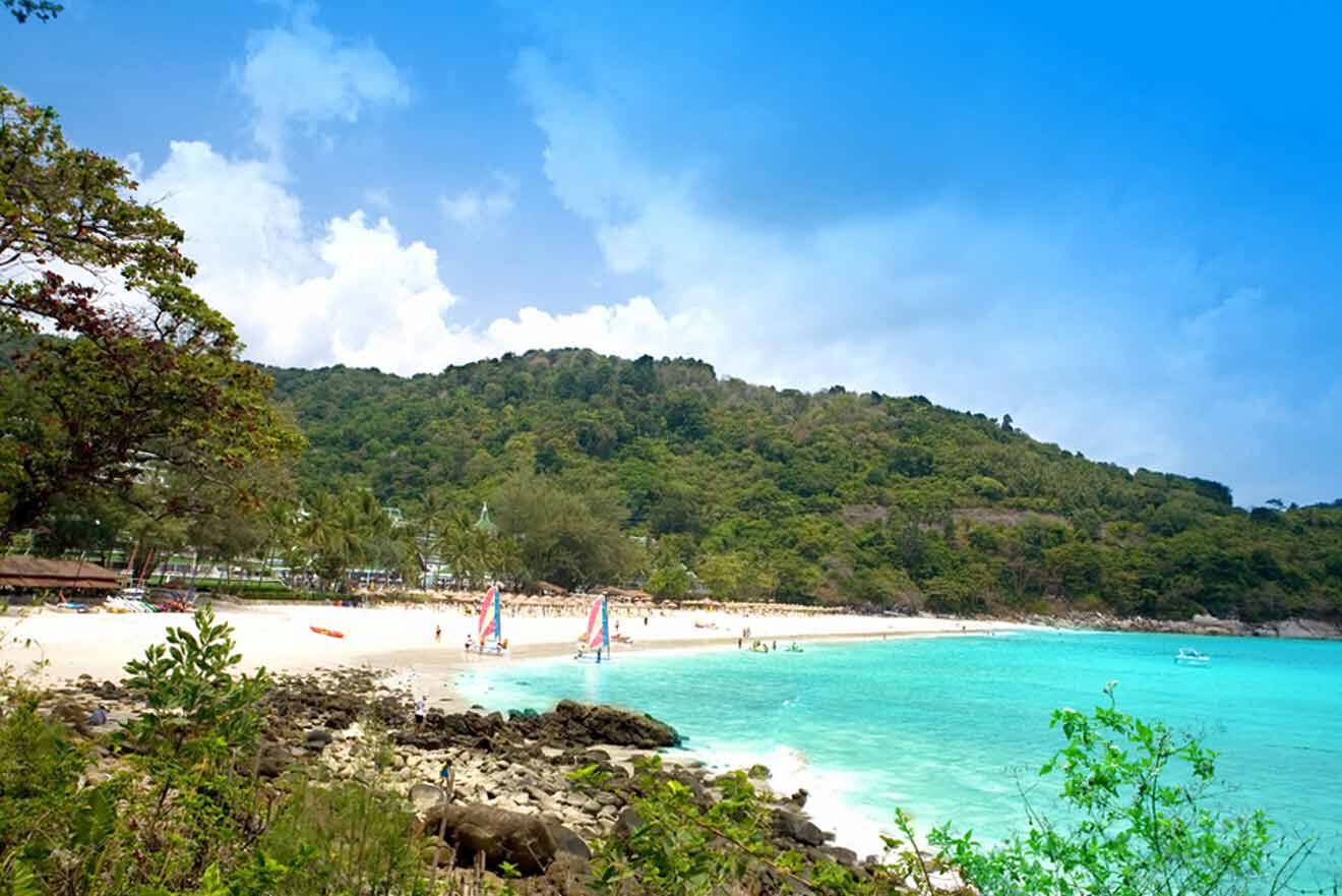 Scenic view of Karon Beach in Thailand, featuring powder-white sand, crystal-clear turquoise waters, and windsurfers enjoying the idyllic beach setting against a backdrop of lush green hills under a blue sky.