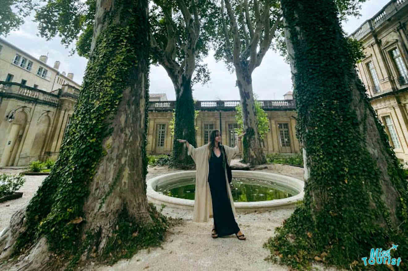 Victoria, the writer of the post in a flowing black dress poses beneath tall trees by a small circular fountain at the Musée Calvet in Avignon, France