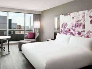 A hotel room with a large comfortable bed, a floral artwork above the headboard, and a window showcasing a view of the city skyline.