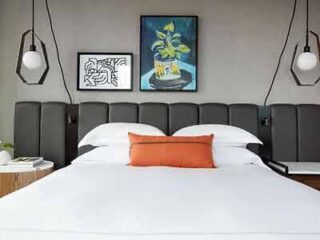 A stylish hotel bedroom featuring a plush headboard, contemporary bedside pendant lights, and a vibrant pop of orange from a throw pillow.