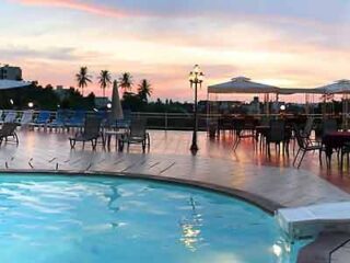Hotel swimming pool at sunset with the sky painted in soft hues of pink and orange, complementing the gentle lighting of the pool area and creating a tranquil evening atmosphere.