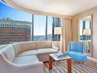 A hotel lounge with panoramic windows offering a city view, featuring a curved sofa and a blue accent chair.