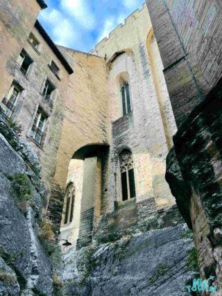 Looking up at the imposing stone architecture of Avignon, France, with Gothic windows and buttresses set against a narrow alley,