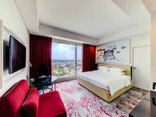 A contemporary hotel room with a large bed, abstract wall art, a red armchair, and a city view from the window.