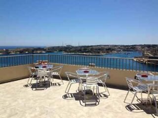 The same sunny hotel rooftop terrace as in image 2, with a closer view of the dining tables and panoramic water vistas.