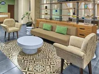 Hotel lobby with modern decor including beige armchairs, a long sofa with green cushions, patterned rug, and a wooden bookshelf.
