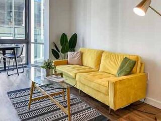 A bright living space with a bold yellow sofa, a geometric rug, and a chic glass coffee table, illuminated by natural light from the large window.