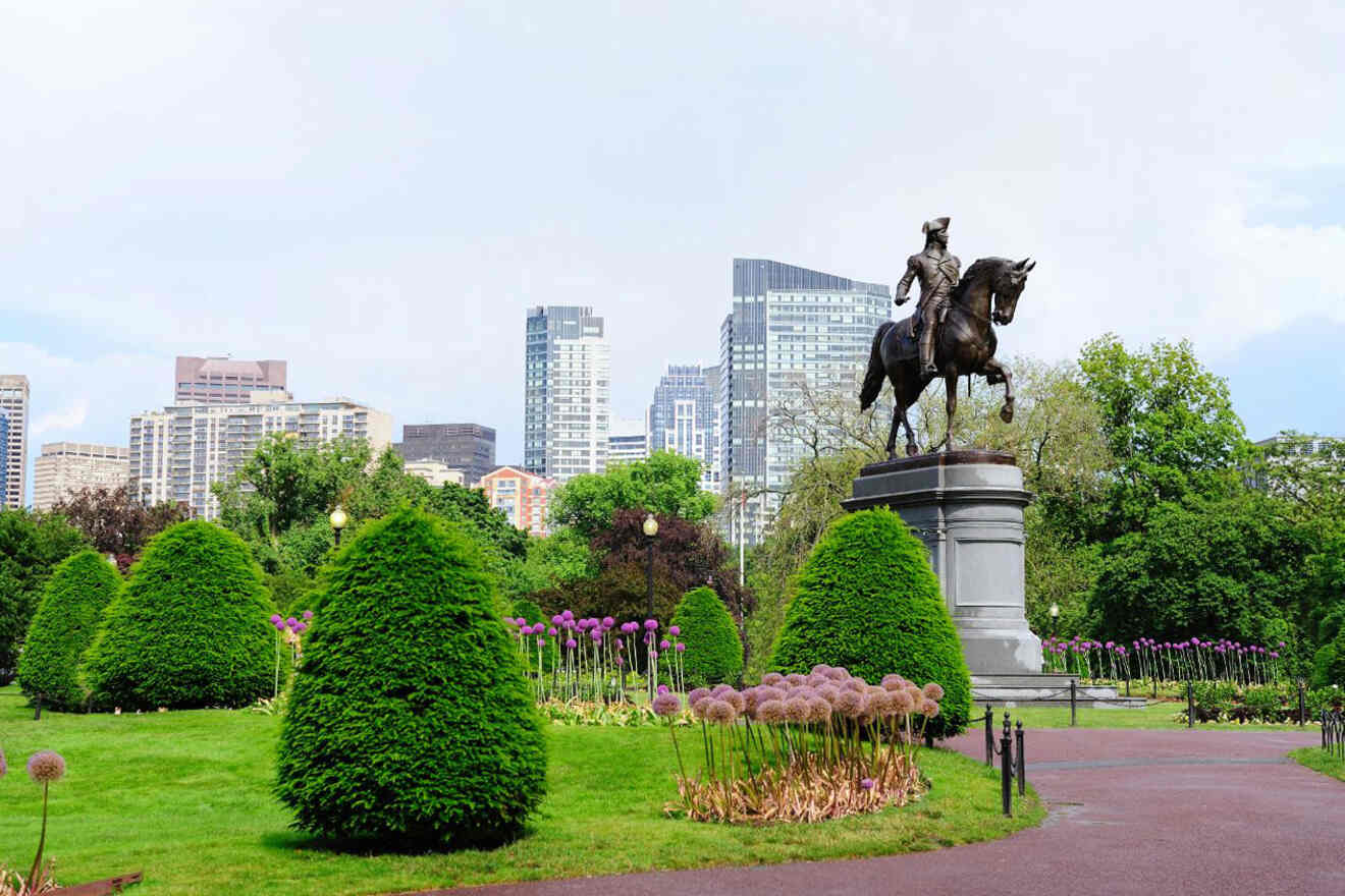 Equestrian statue of a historical figure in the Boston Public Garden with neatly trimmed green shrubs and a backdrop of city buildings under a blue sky