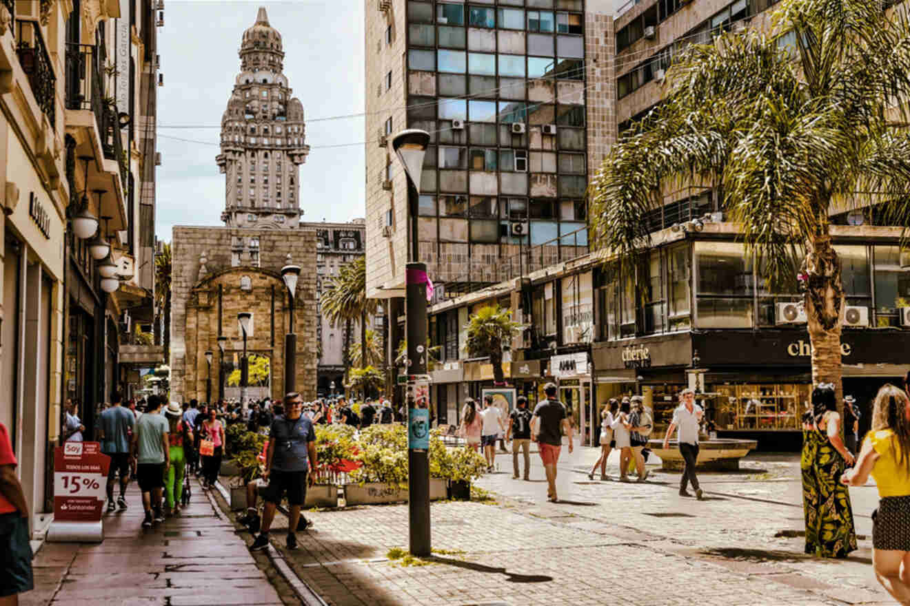 Busy pedestrian street bustling with activity in Montevideo, leading towards the distinctive architecture of Palacio Salvo under a clear sky, with local shops lining the sidewalk and a variety of people walking and enjoying the urban atmosphere.