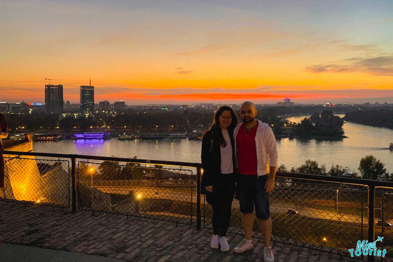 The writer of the post with her partner poses at twilight with a vibrant sunset over the Belgrade skyline and river, with the city's lights beginning to twinkle