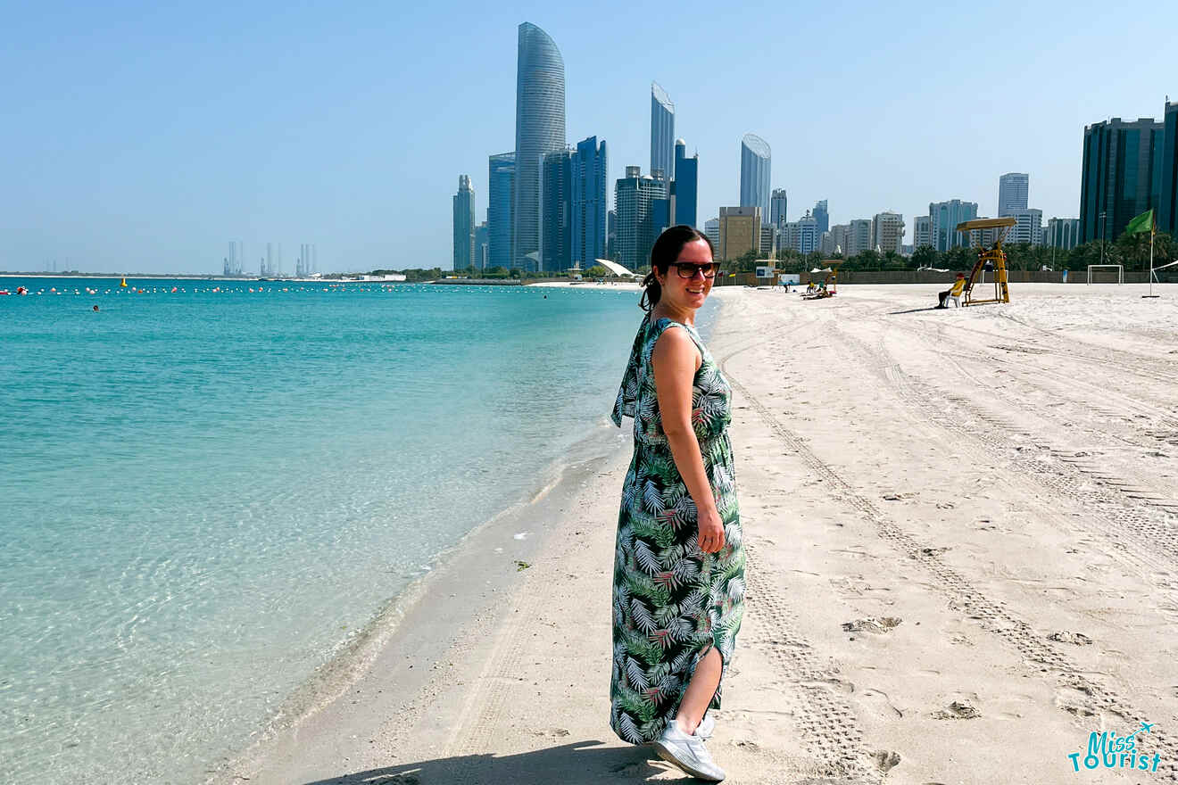 Writer of the post  in a tropical print dress smiles at the camera on the sandy Corniche Beach with the Abu Dhabi skyline in the background under a clear blue sky, with text 'Miss Tourist' in the corner.