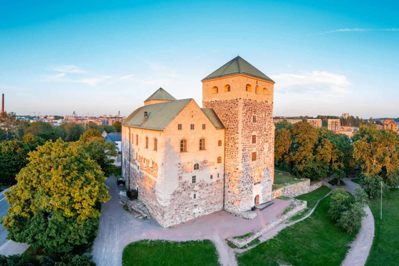 Aerial view of the historic Turku Castle at dusk, surrounded by lush greenery with the cityscape in the background