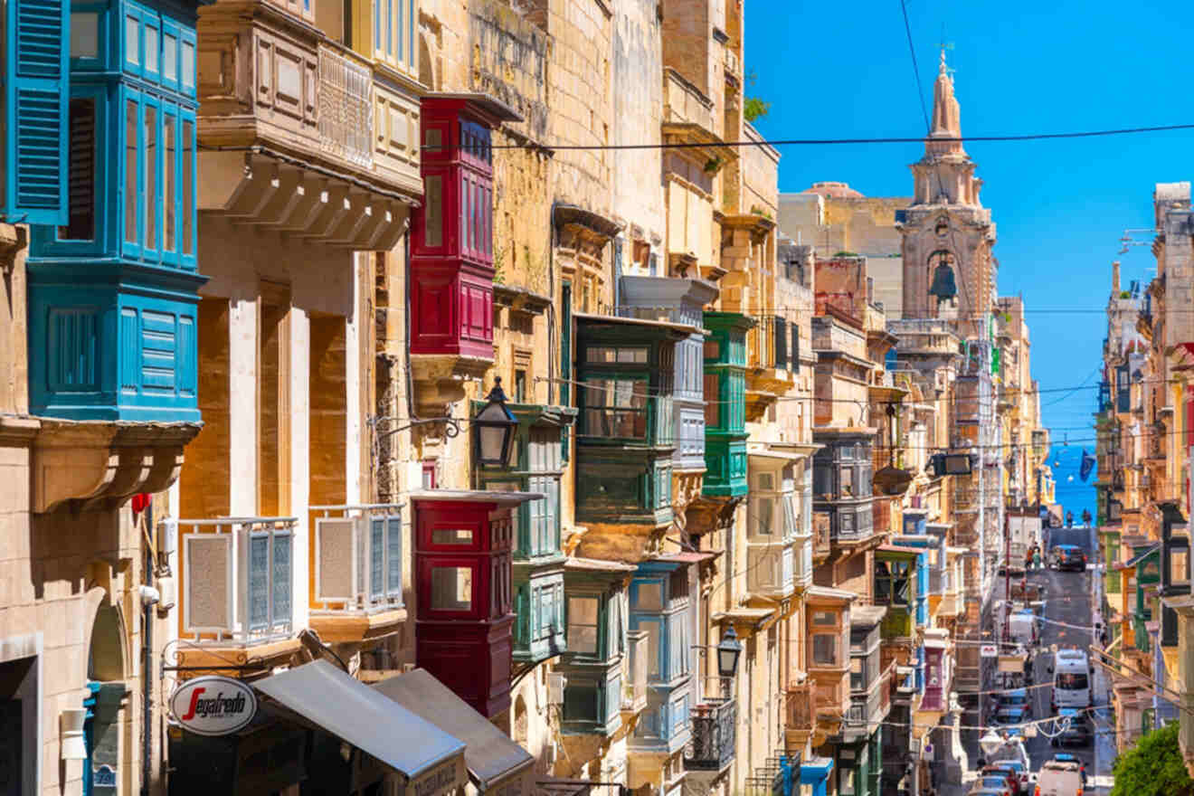 Colorful traditional Maltese balconies lining a steep street in Valletta, leading down to the sea, with the baroque architecture of St. Paul's Pro-Cathedral in the background.