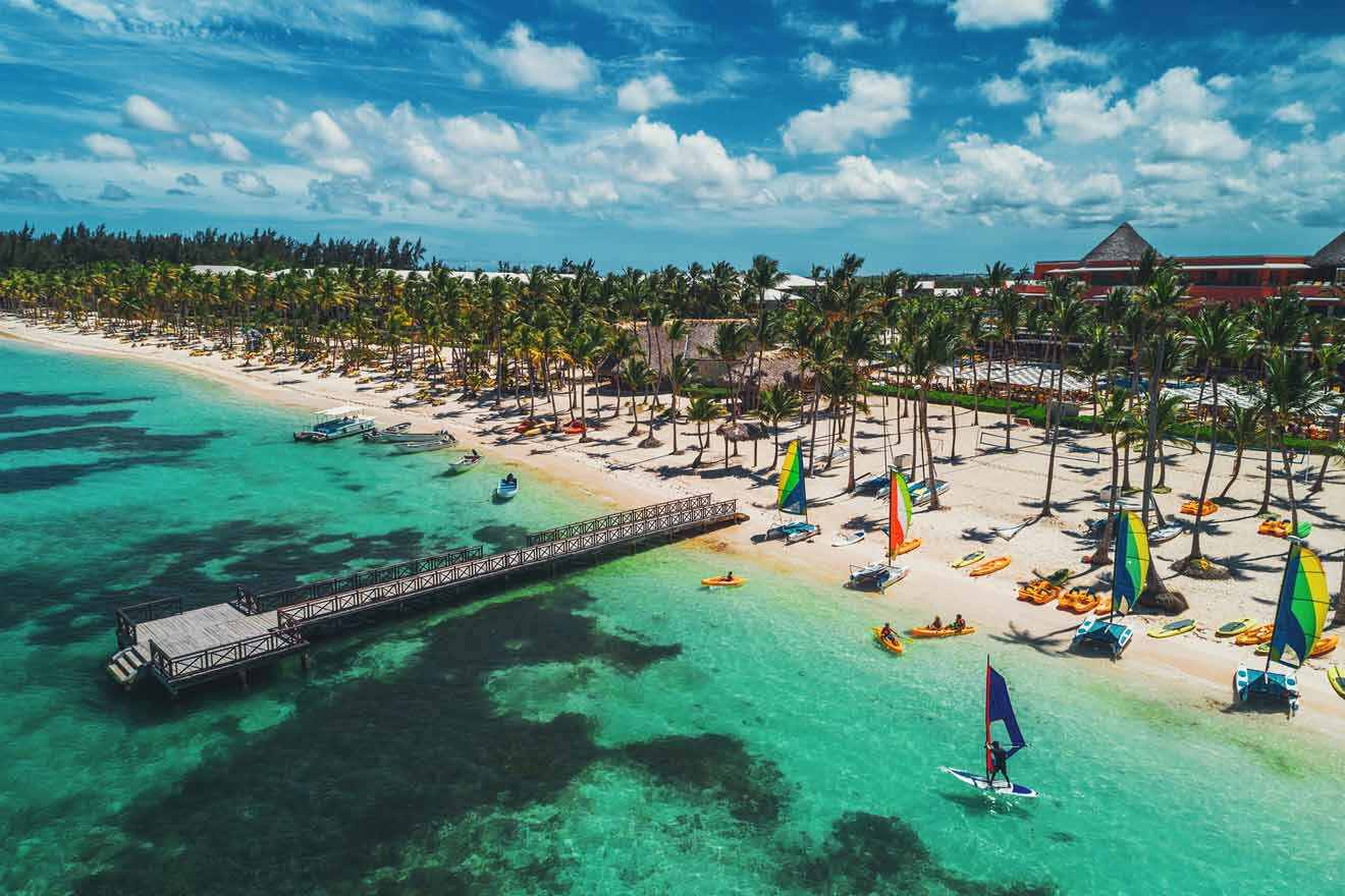 Aerial view of Bavaro Beach in Punta Cana, featuring crystal clear turquoise waters, colorful sailboats, a wooden pier, and a line of palm trees against a sunny sky.